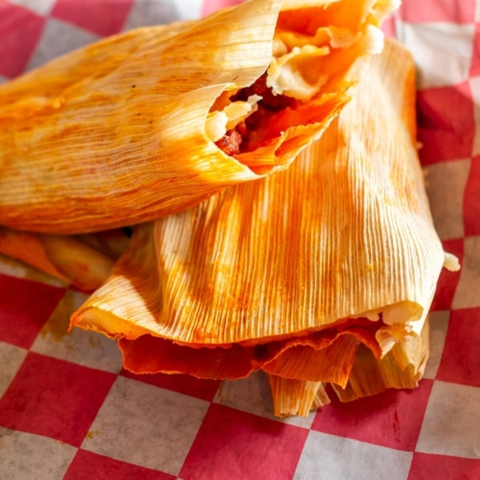 Two tamales with red filling, partially unwrapped, resting on red and white checkered paper.