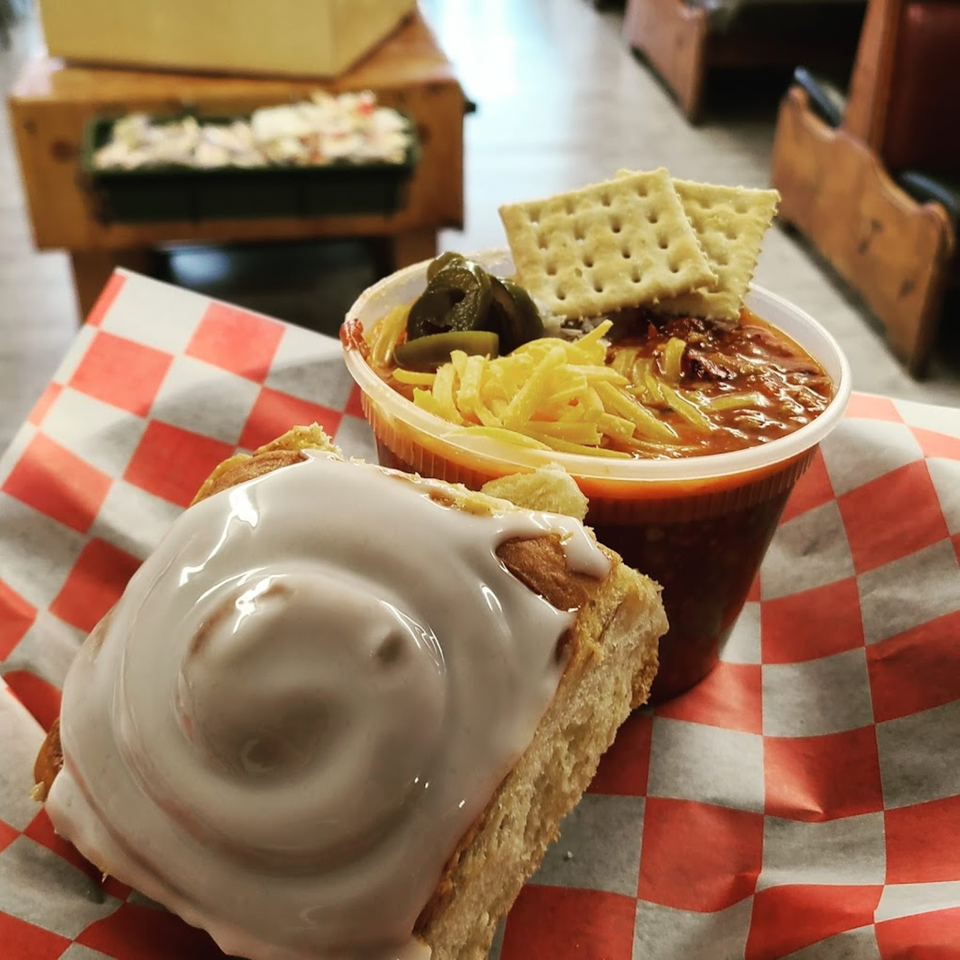 Chili with cheese, jalapeños, and crackers, next to a cinnamon roll on red checkered paper.