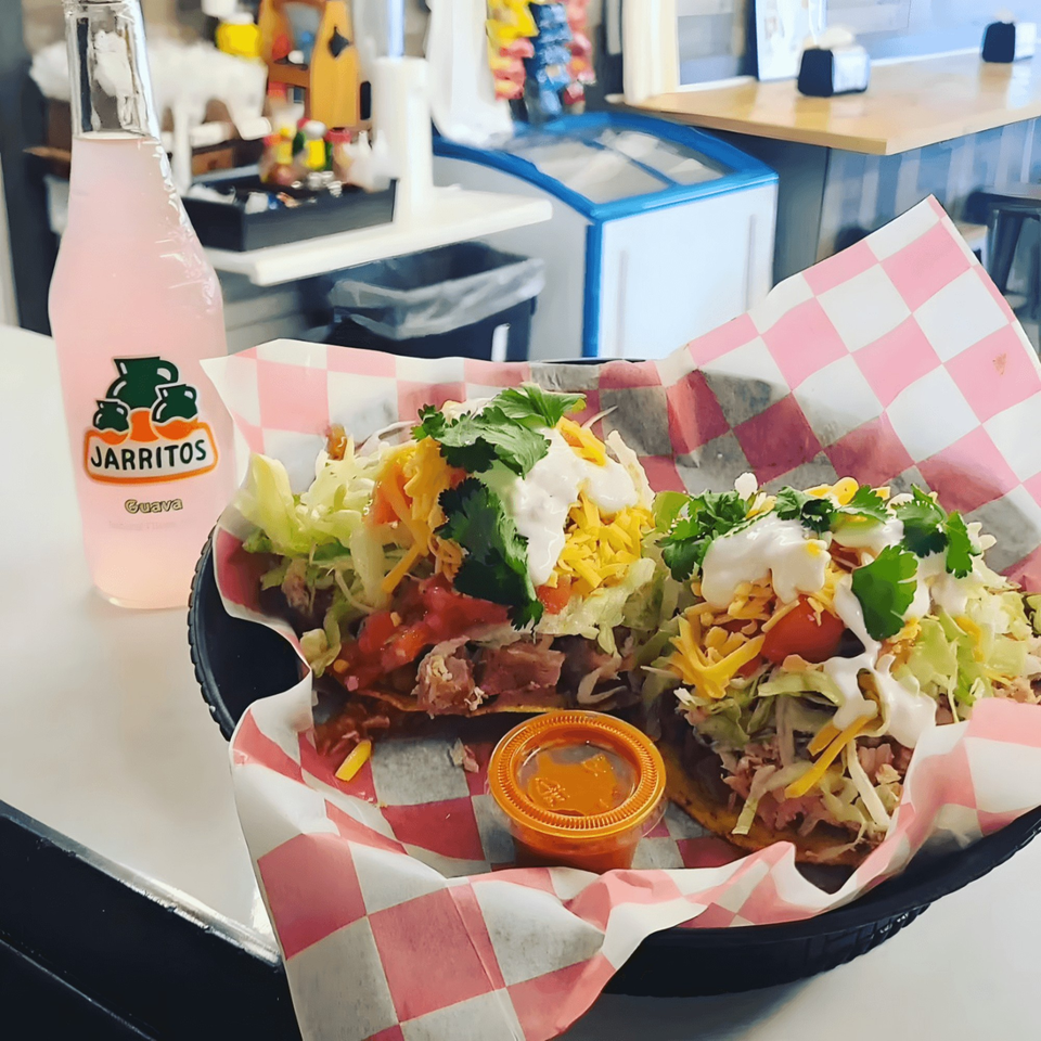 Two loaded tacos with lettuce, cheese, sour cream, and cilantro, next to a pink Jarritos soda.