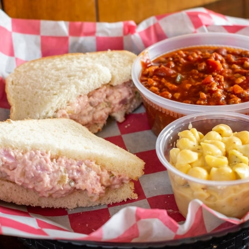 Two sandwiches with pink filling, chili, and macaroni and cheese in a red and white checkered basket.