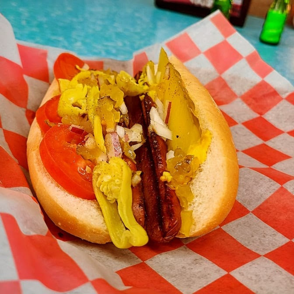 A Chicago-style hot dog with tomatoes, pickles, relish, and peppers in a red-checkered paper.